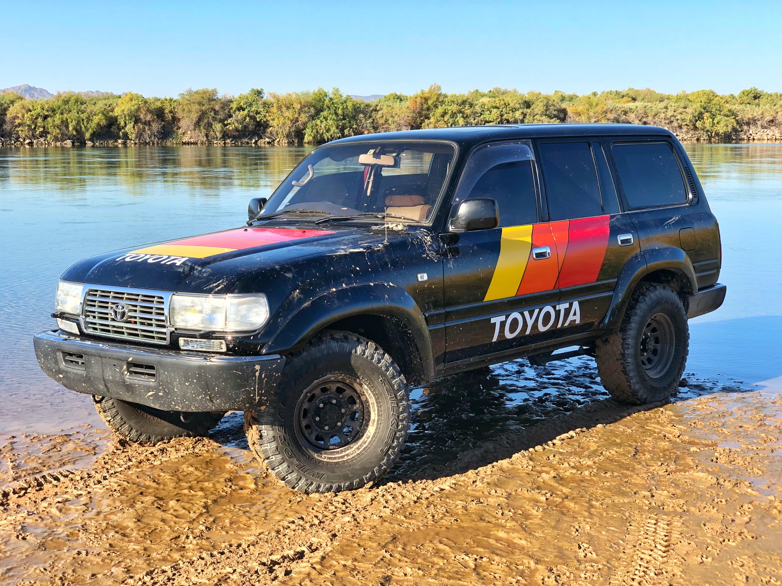 Black 80 Series Land Cruiser with retro Toyota racing stripes on a riverbank